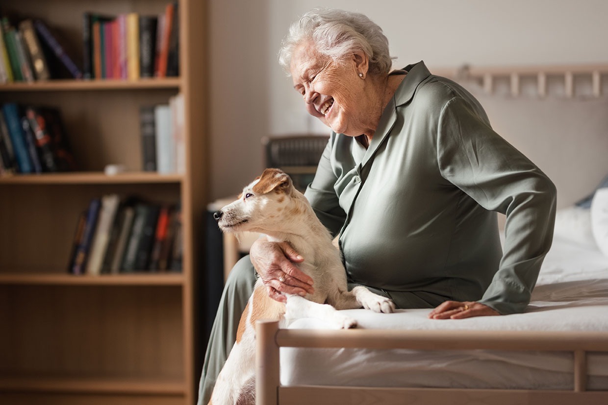 An elderly woman in an aged care home holding a dog with a smile.